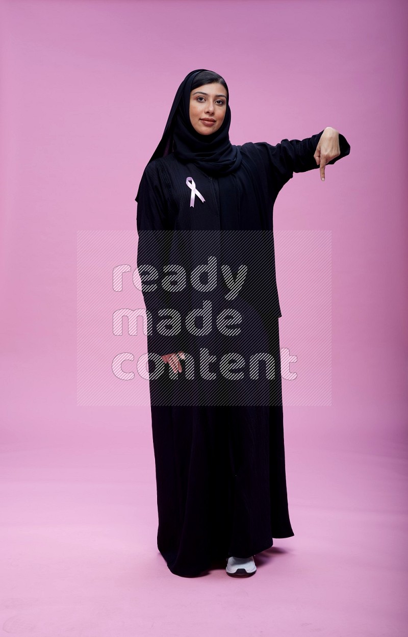 Saudi woman wearing pink ribbon on Abaya standing interacting with the camera on pink background