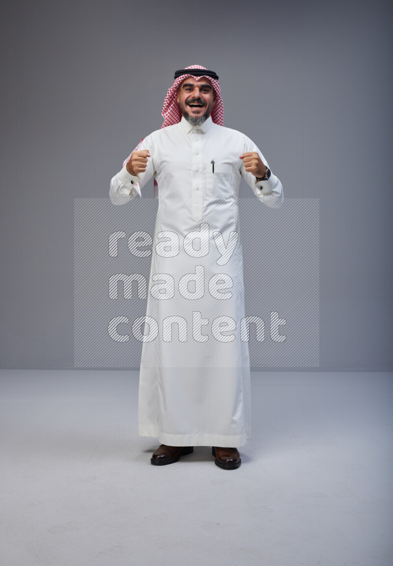Saudi man Wearing Thob and red white Shomag standing interacting with the camera on Gray background