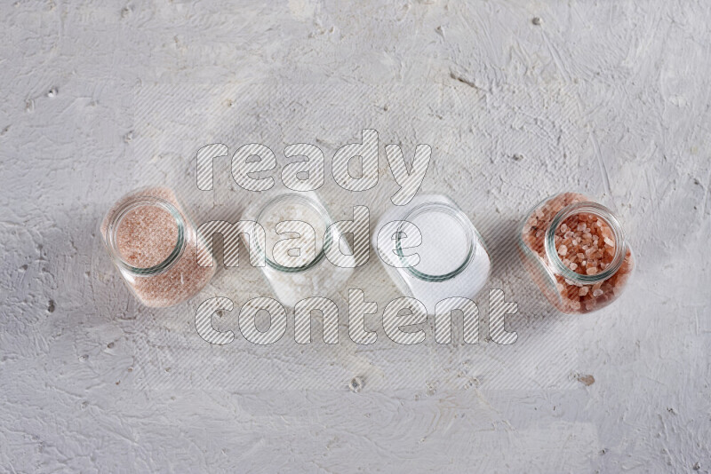 4 glass jars filled with table salt, coarse sea salt, fine himalayan salt and coarse himalayan salt on white background