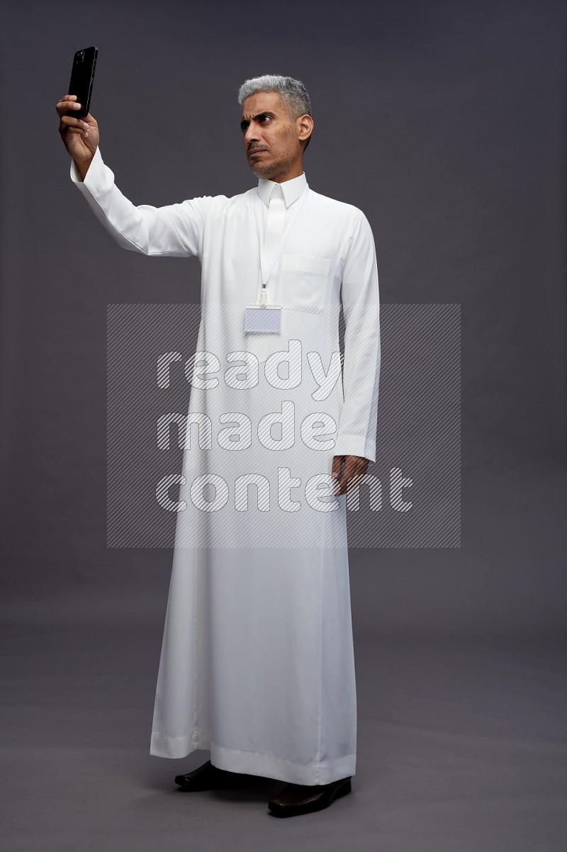 Saudi man wearing thob with neck strap employee badge standing taking selife on gray background