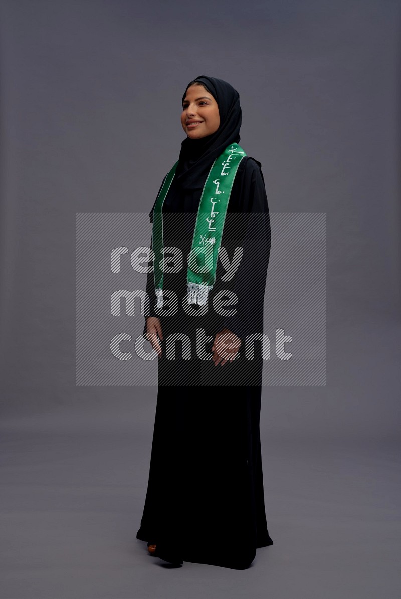 Saudi woman wearing Abaya standing holding Saudi flag on gray background