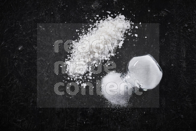 A glass jar full of table salt with some sea salt crystals beside it on a black background