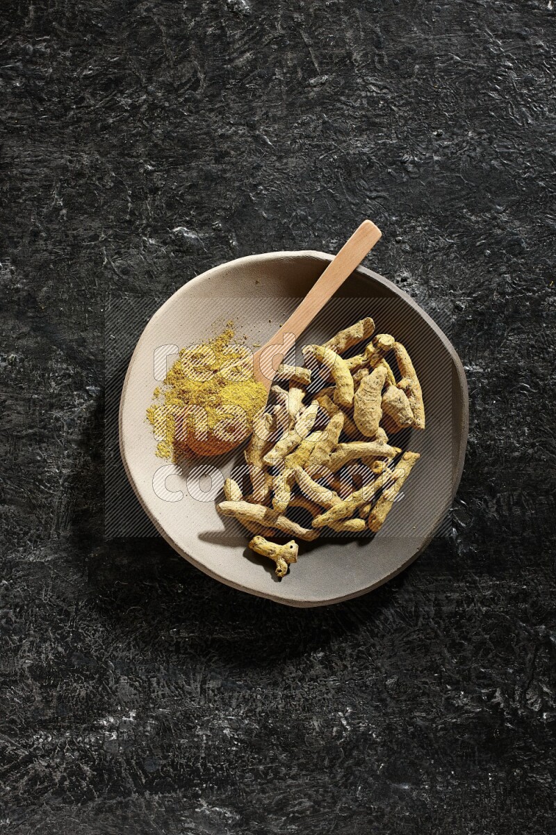 A plate filled with dried turmeric fingers and a wooden spoon full of turmeric powder on a textured black flooring