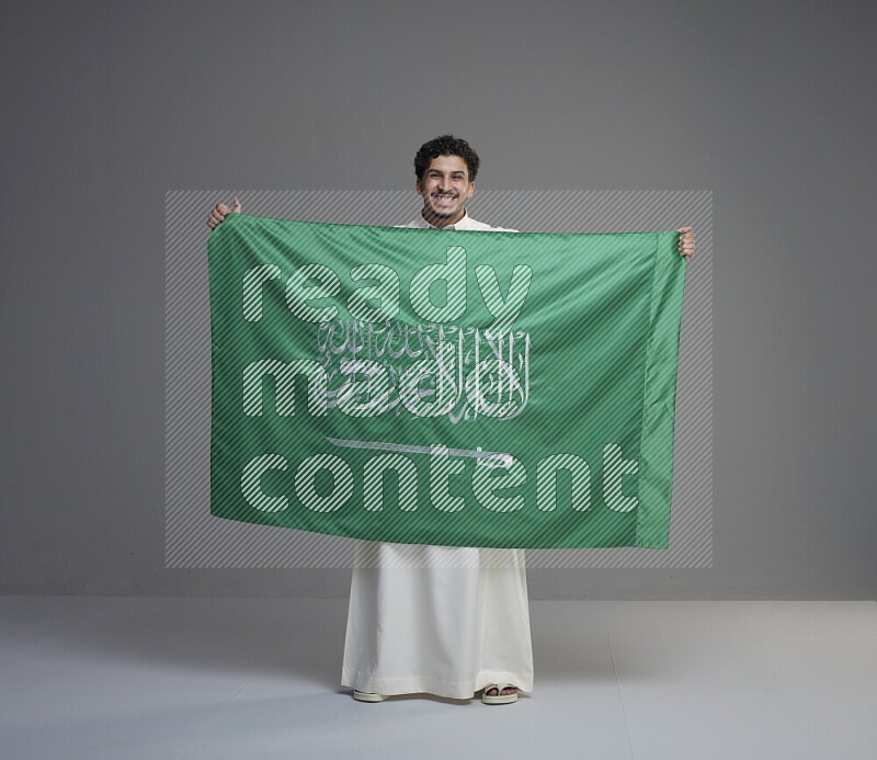 A saudi man standing wearing thob holding big saudi flag on gray background