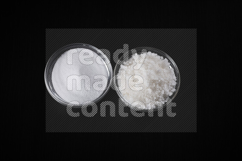 2 glass bowls one is filled with table salt and the other with coarse sea salt on black background