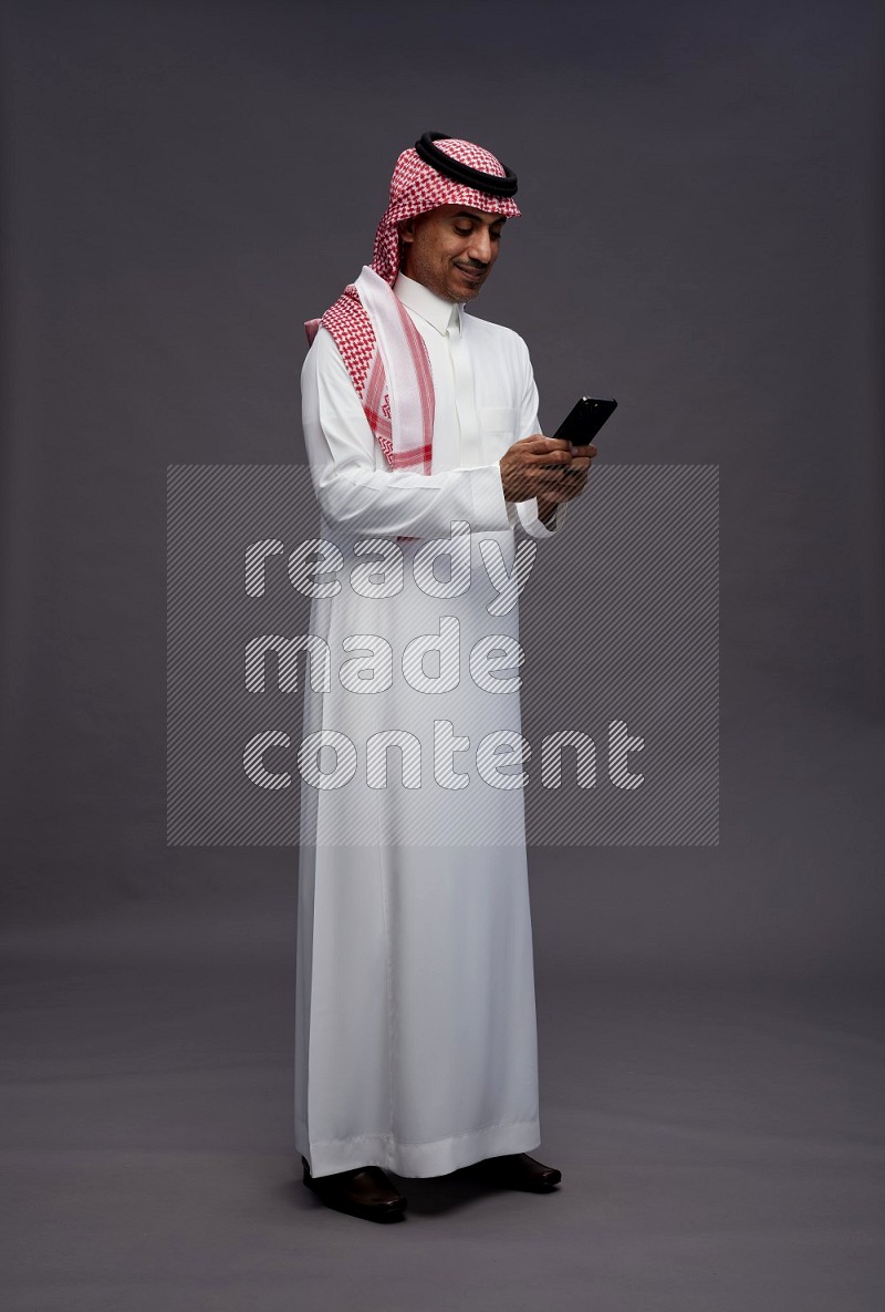 Saudi man wearing thob and shomag standing texting on phone on gray background
