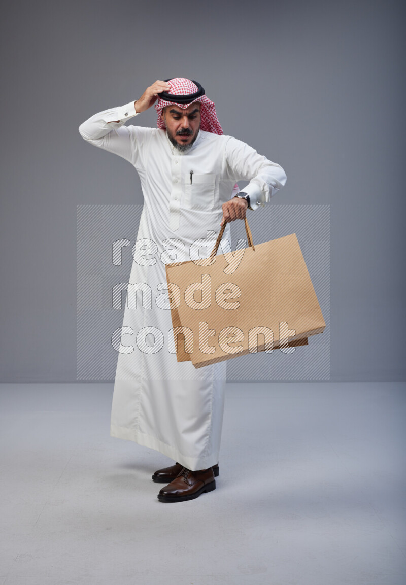 Saudi man Wearing Thob and red Shomag standing holding shopping bag on Gray background