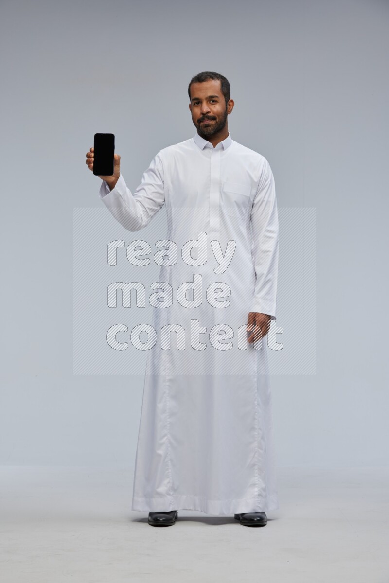 Saudi man wearing Thob standing showing phone to camera on Gray background