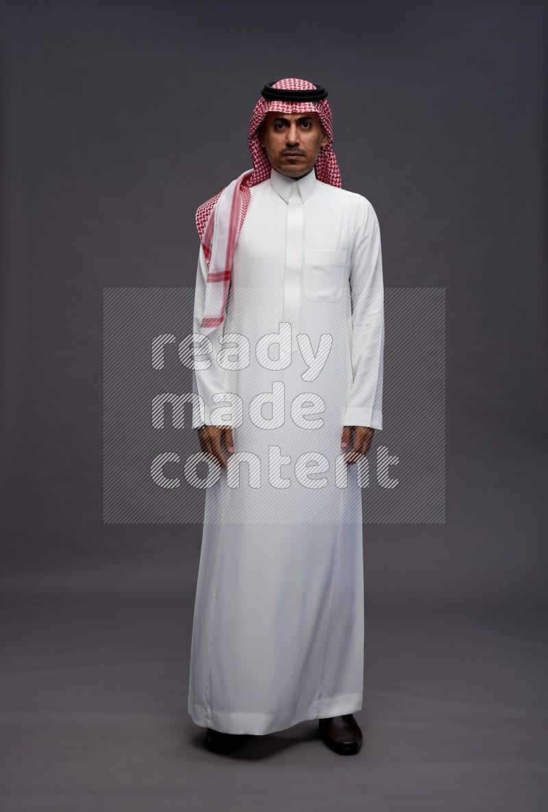 Saudi man wearing thob and shomag standing interacting with the camera on gray background