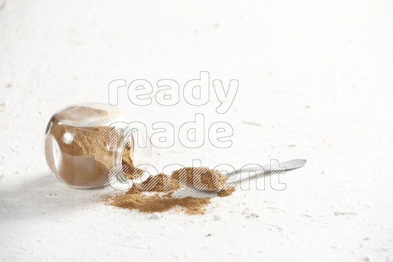 Flipped herbs glass jar full of cinnamon powder with a metal spoon full of powder on a textured white background