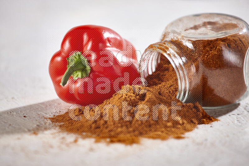 A glass jar full of ground paprika powder flipped with some spilling powder on white background