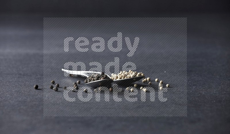 2 metal spoons full of black and white pepper beads with spreaded beads on black flooring