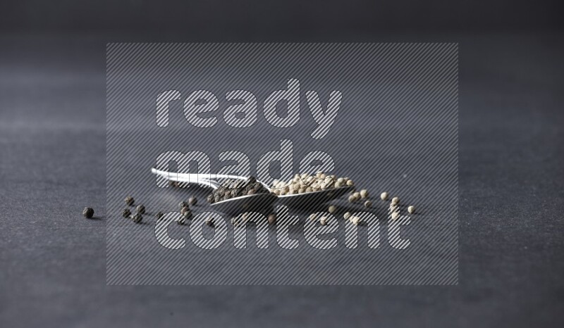 2 metal spoons full of black and white pepper beads with spreaded beads on black flooring