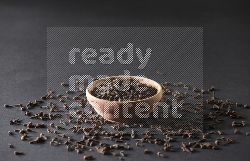 A wooden bowl full of cloves with spreaded whole cloves on a black flooring