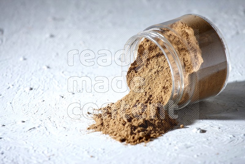 A flipped glass jar full of allspice powder and powder spilled out of it on a textured white flooring