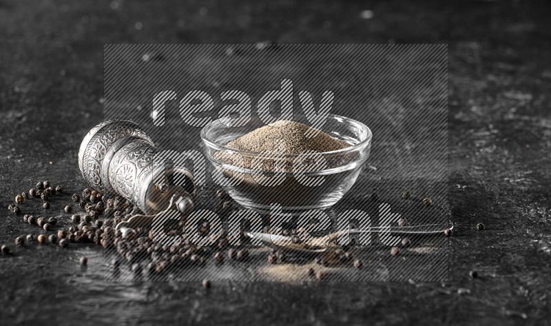A glass bowl full of black pepper powder with black pepper beads, a turkish metal pepper grinder and a metal spoon on textured black flooring