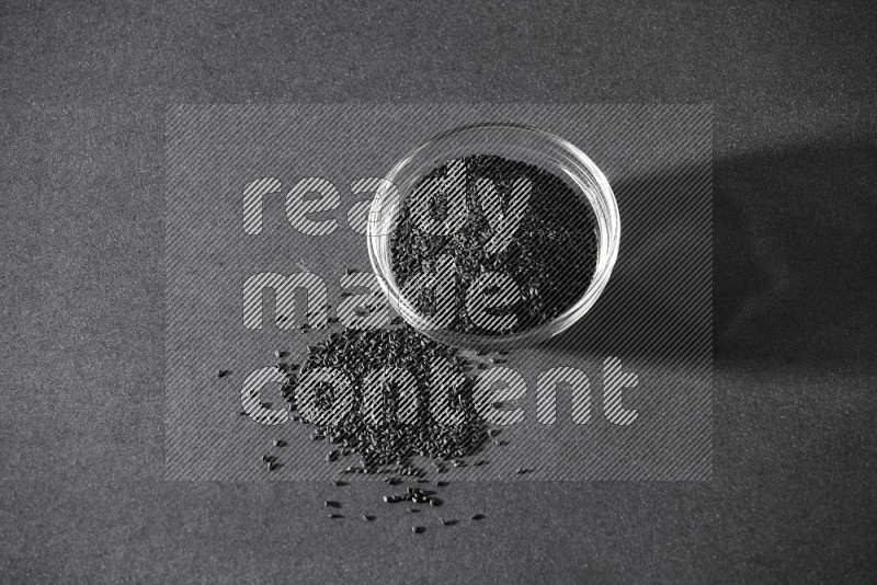A glass bowl full of black seeds and seeds spreaded beside it on a black flooring in different angles