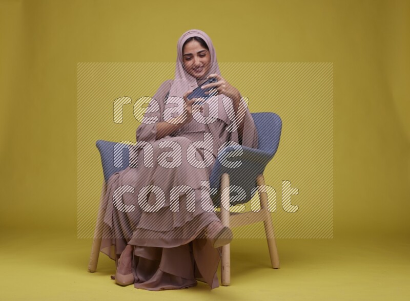 A woman Playing Game Sitting  on a Yellow Background wearing Brown Abaya with Hijab