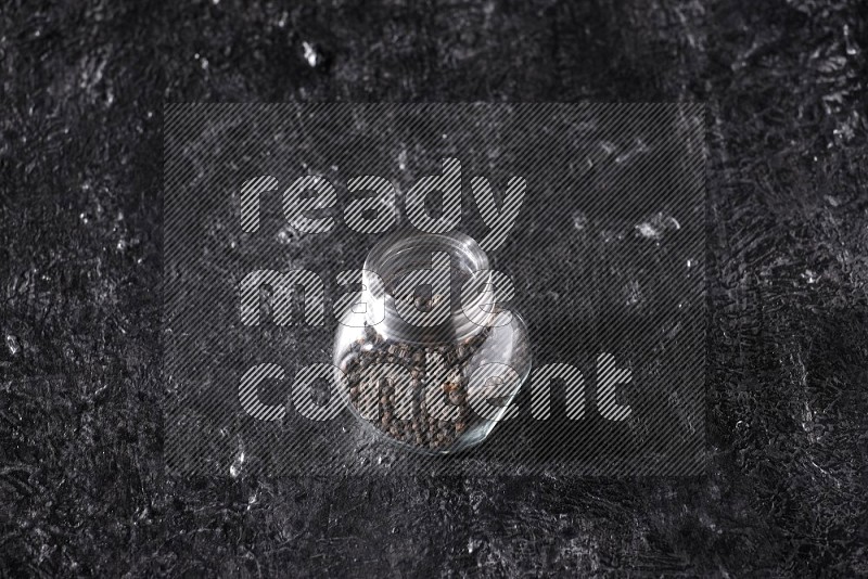 A glass spice jar full of black pepper beads on textured black flooring