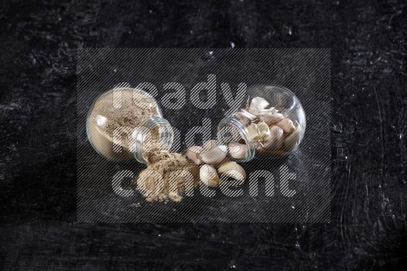 2 glass spice jars full of garlic cloves and powder flipped and the garlic came out on a textured black flooring in different angles