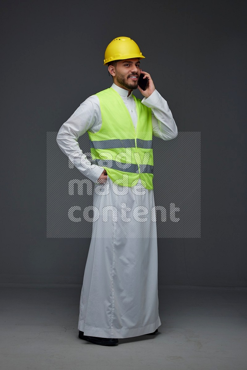 A Saudi man wearing Thobe with a yellow safety vest and white helmet standing and talking in the phone eye level on a gray background