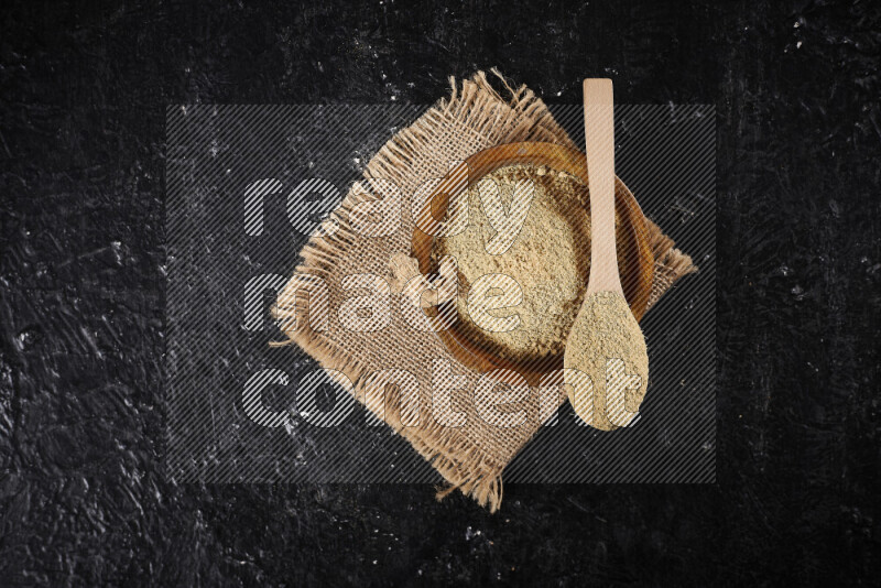 A wooden bowl full of ground ginger powder with a wooden spoon on it all on a burlap fabric on black background