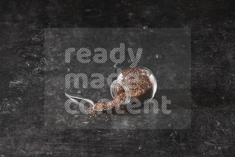 A glass spice jar full of flaxseeds flipped and seeds spread out with a metal spoon full of the seeds on a textured black flooring