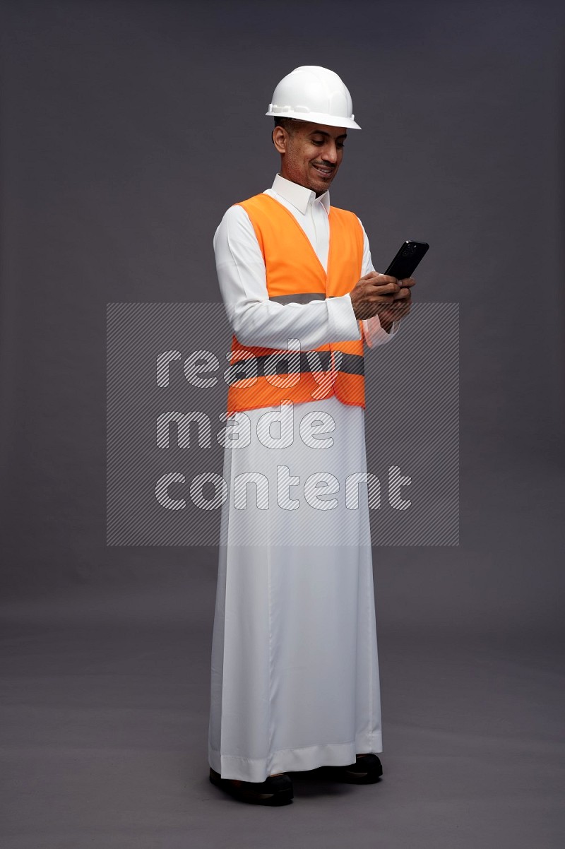 Saudi man wearing thob with engineer vest standing texting on phone on gray background