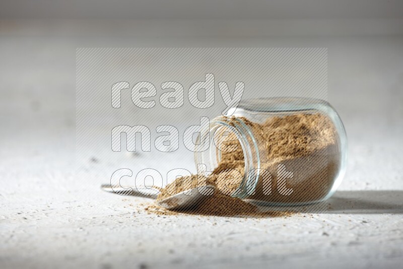 A flipped glass spice jar and a metal spoon full of cumin powder and powder spilled out on textured white flooring