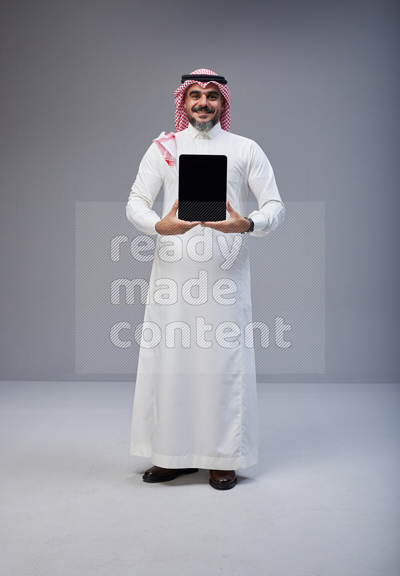 Saudi man Wearing Thob and red Shomag standing showing tablet to camera on Gray background