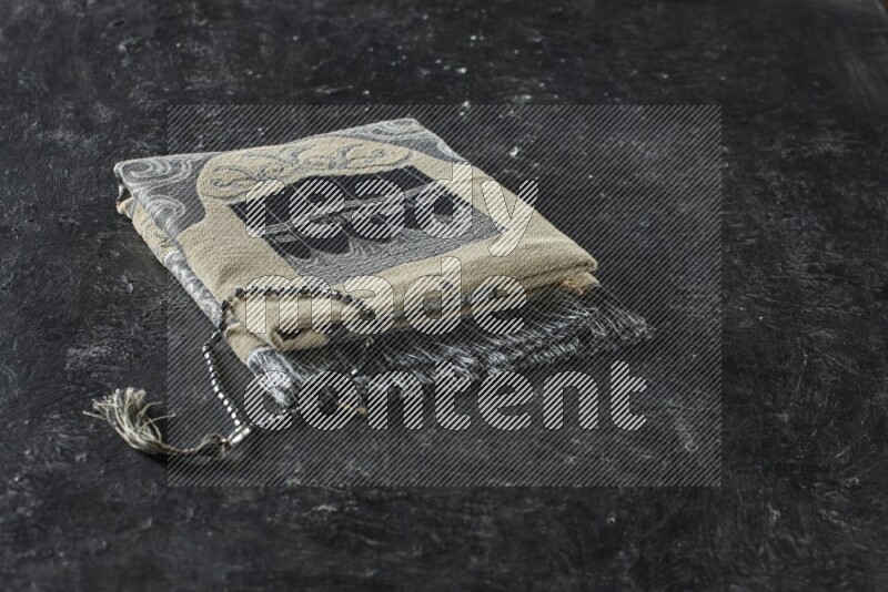 A folded prayer mat with different elements such as quran and prayer beads on black textured background