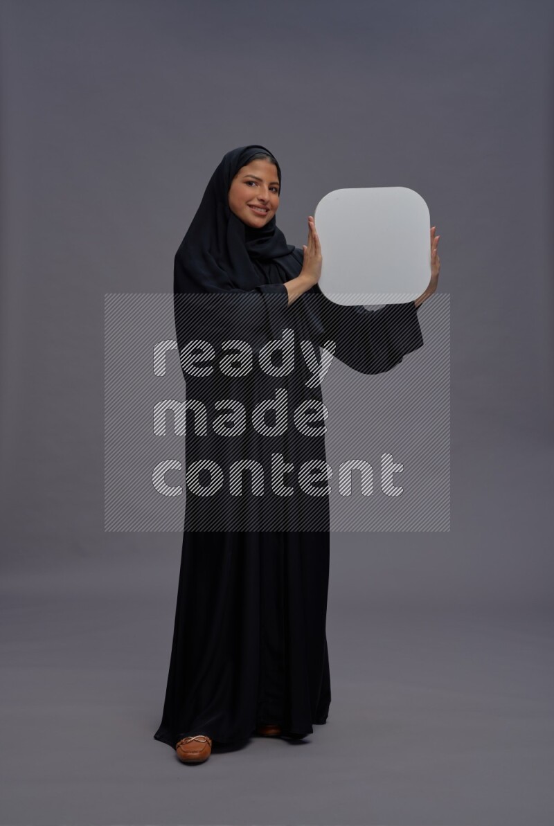 Saudi woman wearing Abaya standing holding social media sign on gray background