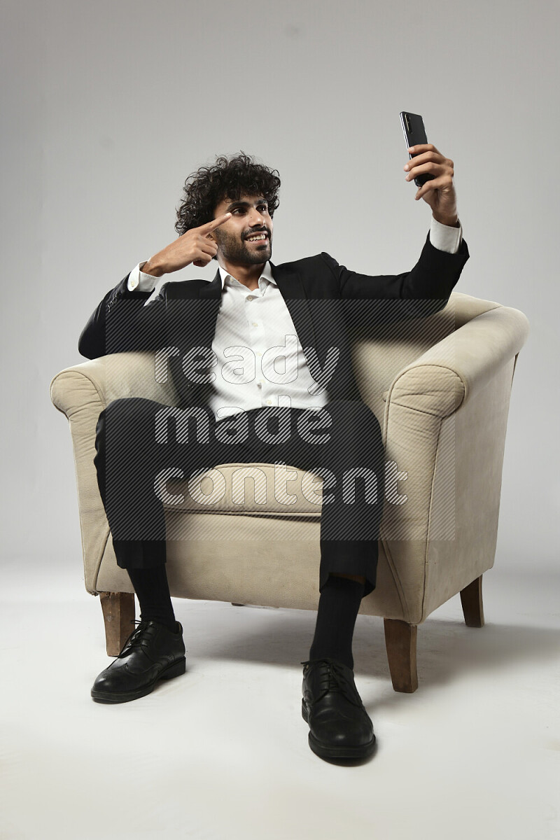 A man wearing formal sitting on a chair taking a selfie on white background