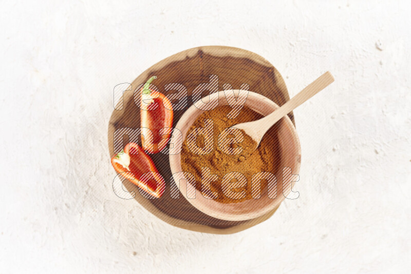 A wooden bowl full of ground paprika powder and sliced red bell pepper beside it all on a wooden tray on white background