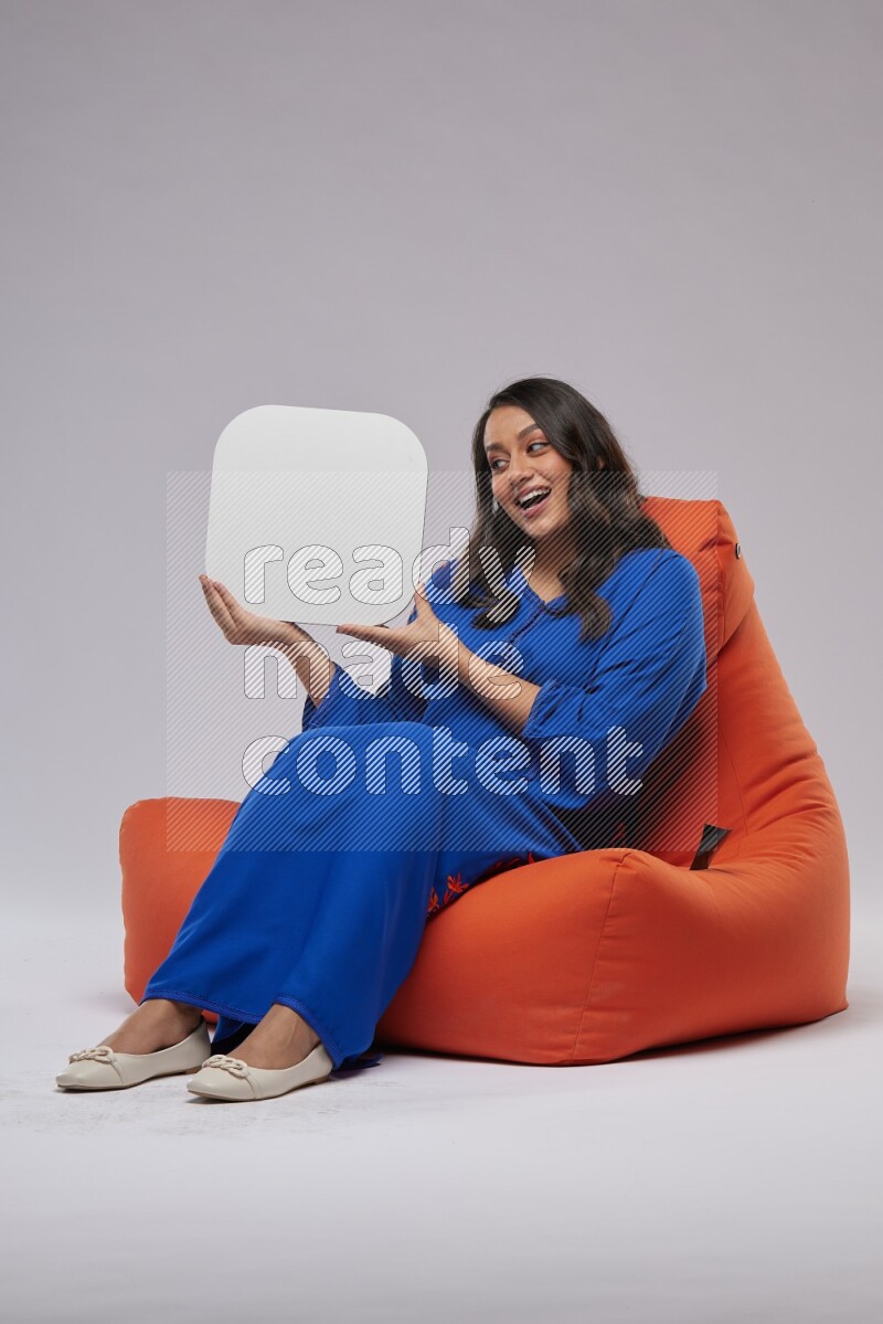 A Woman sitting on an orange beanbag wearing Jalabeya holding a social media sign
