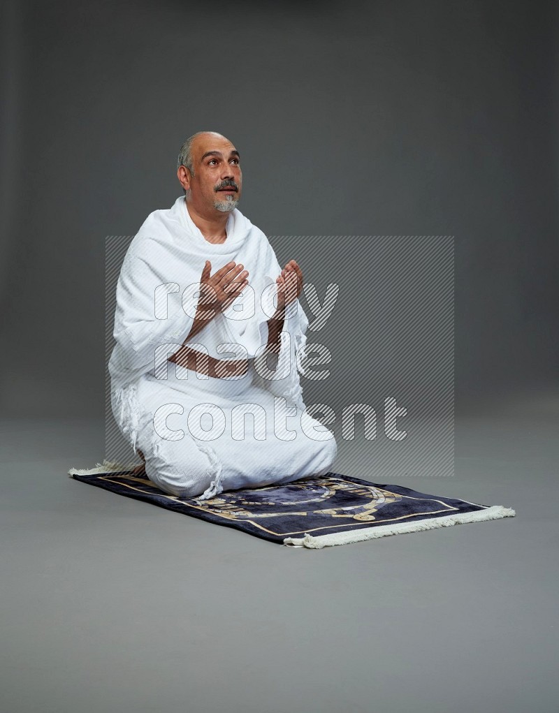 A man wearing Ehram sitting on prayer mat dua'a on gray background