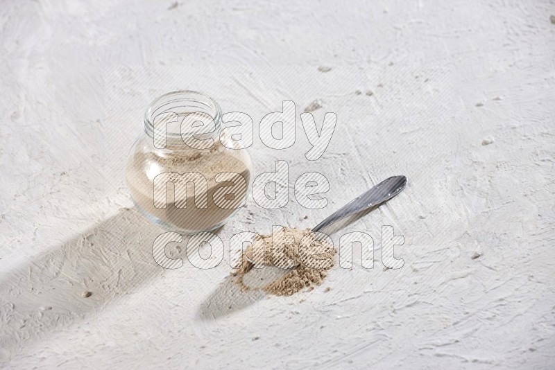 A glass spice jar full of garlic powder with metal spoon on a textured white flooring in different angles