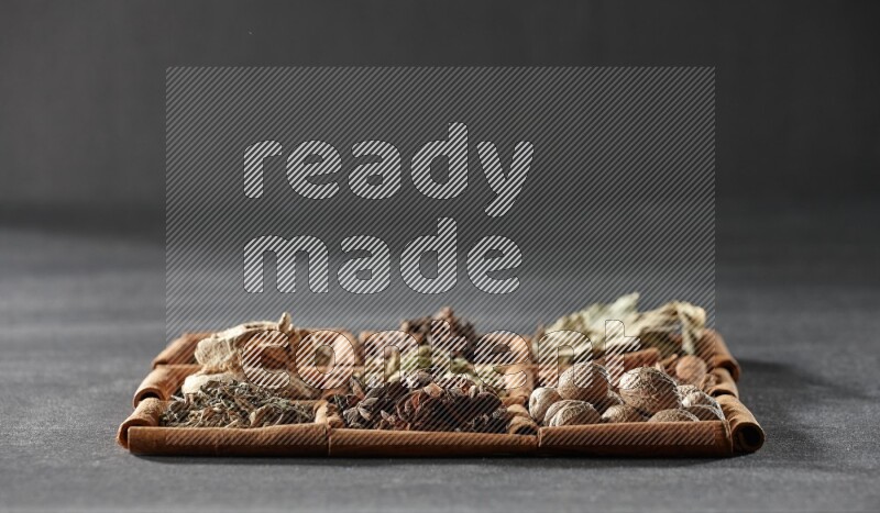 9 مربعات من أعواد القرفة ممتلئة بالهيل في المنتصف محاطة بجوزة الطيب والقرفة وأوراق الغار والقرنفل والكمون والزنجبيل المجفف والريحان المجفف واليانسون على أرضية سوداء