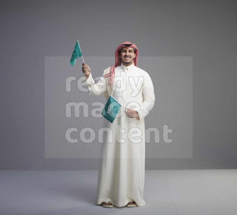 A Saudi man standing wearing thob and red shomag raising small Saudi flag on gray background