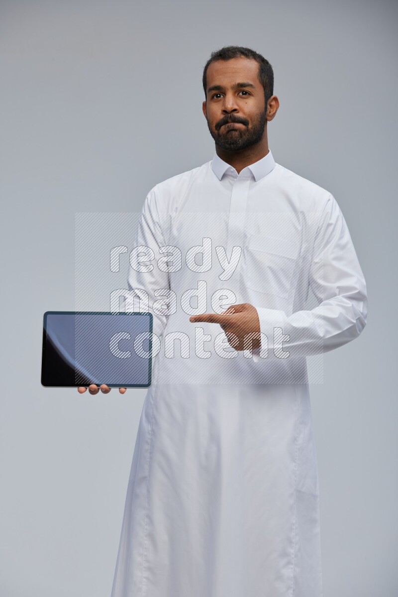 Saudi man wearing Thob standing showing tablet to camera on Gray background