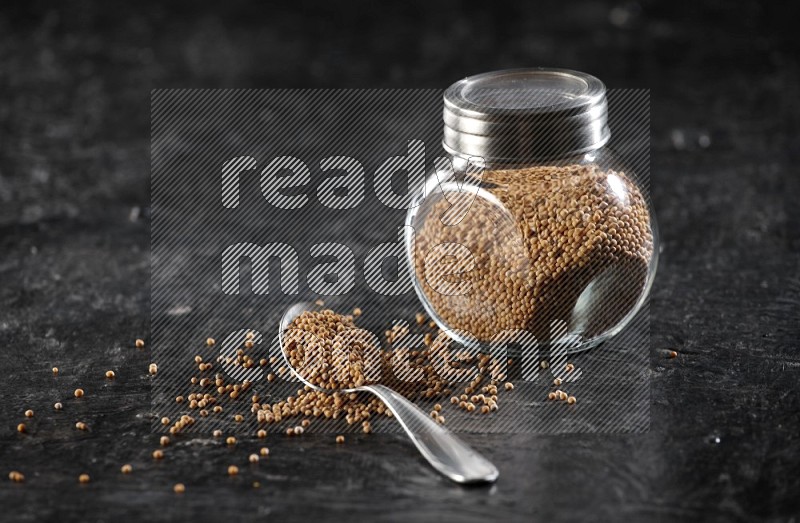 A glass spice jar and a metal spoon full of mustard seeds on a textured black flooring in different angles