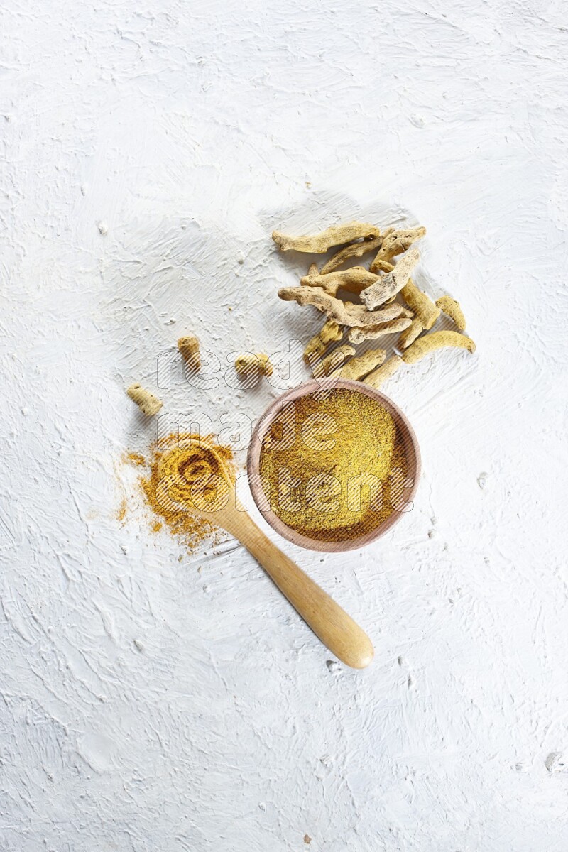 A wooden bowl and wooden spoon full of turmeric powder with dried turmeric fingers beside it on textured white flooring