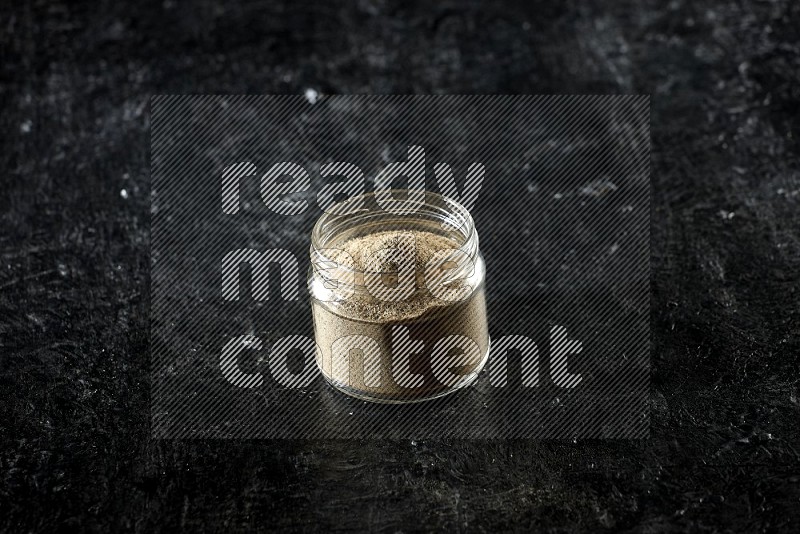 A glass jar full of cardamom powder on textured black flooring