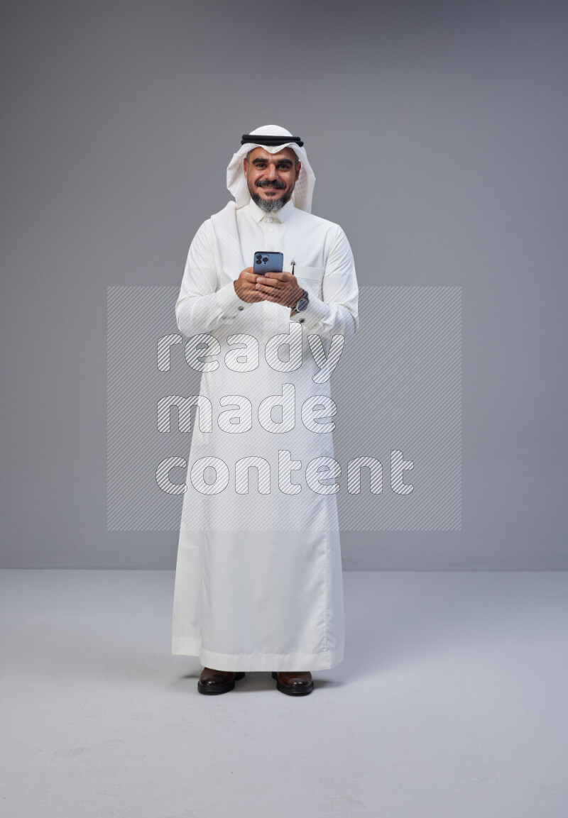 Saudi man Wearing Thob and white Shomag standing texting on phone on Gray background