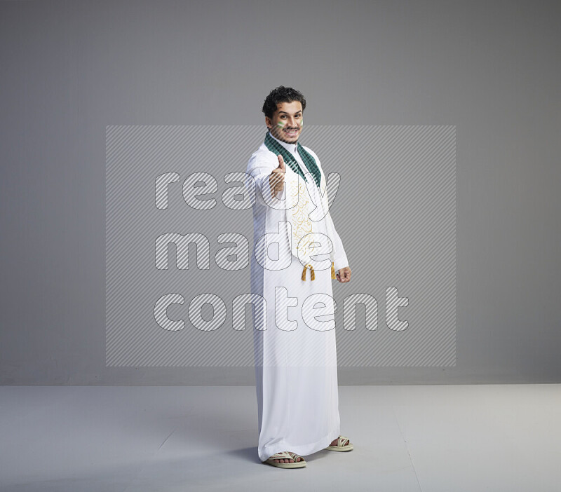 A Saudi man standing wearing thob and Saudi flag scarf with face painting on gray background