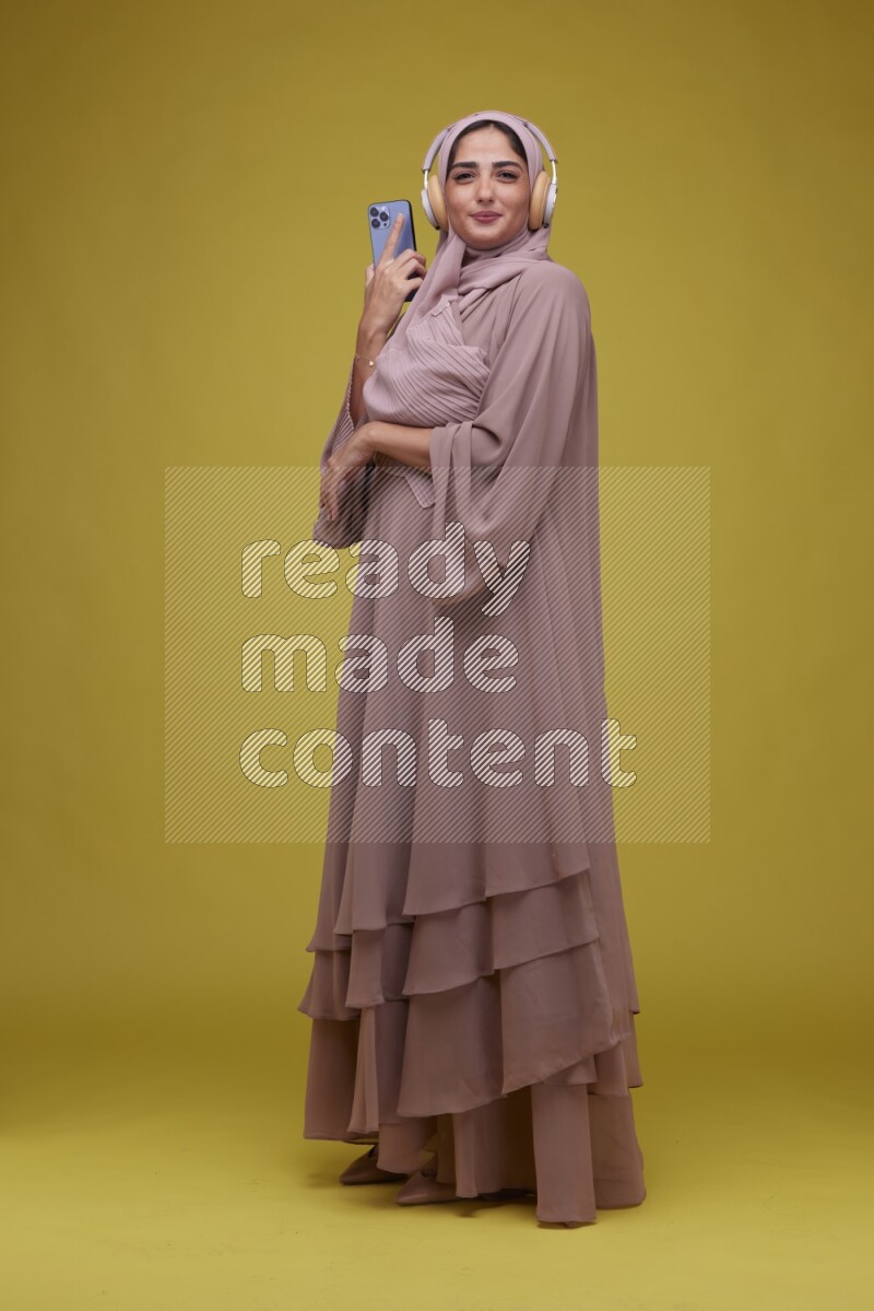 A woman Listening to Music on a Yellow Background wearing Brown Abaya with Hijab