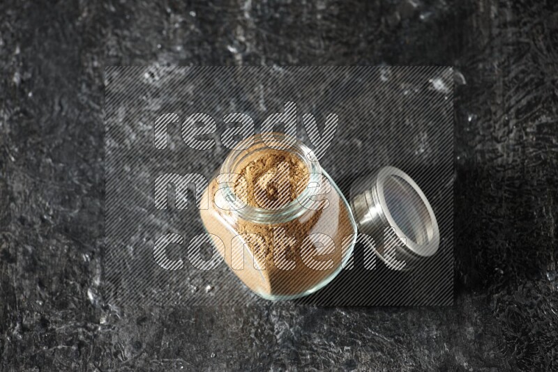 A glass spice jar full of allspice powder on a textured black flooring