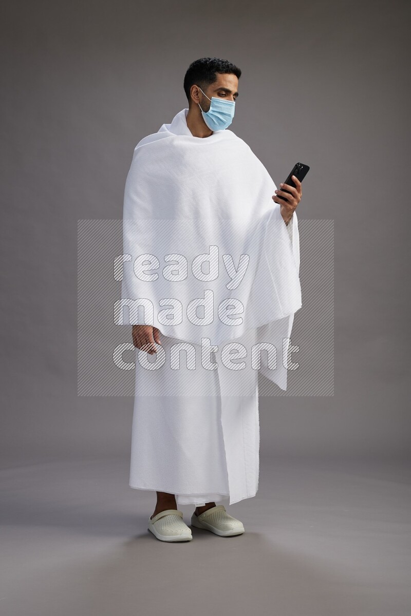 A man wearing Ehram with face mask Standing texting on phone on gray background