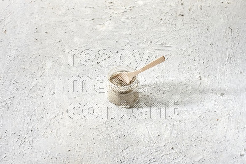 A glass jar and wooden spoon full of white pepper powder on textured white flooring