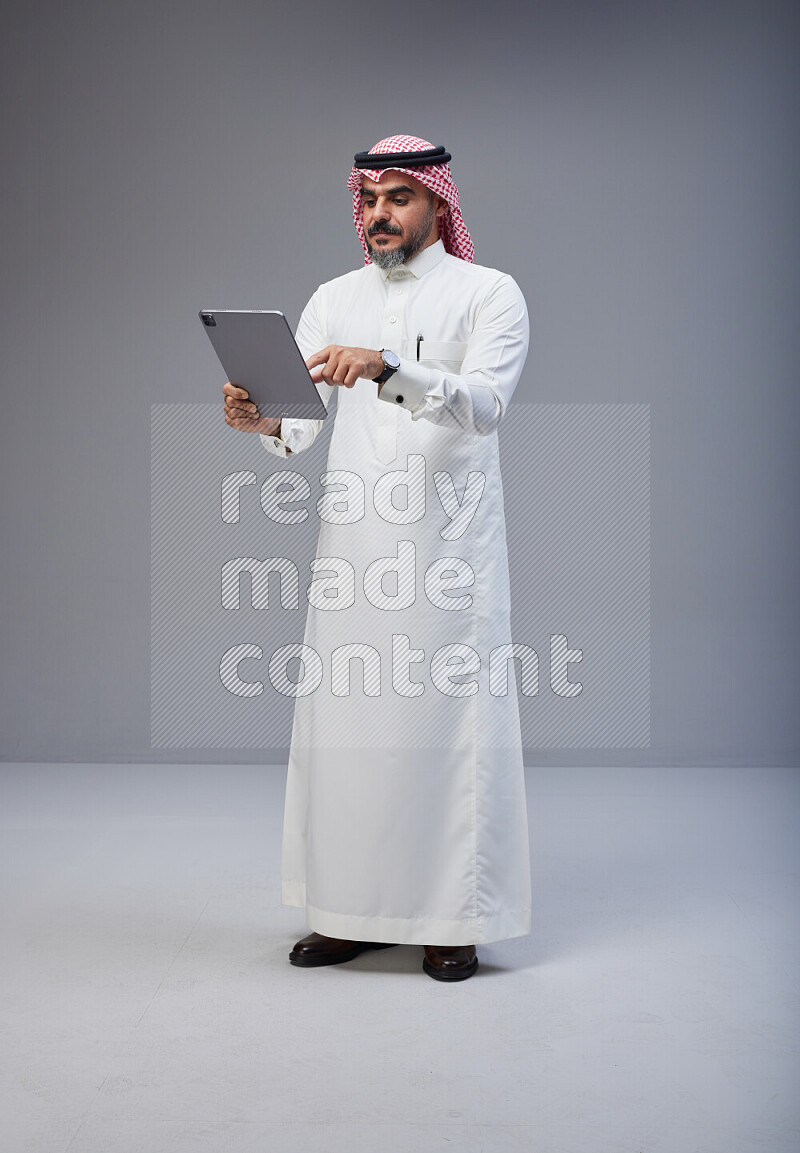 Saudi man Wearing Thob and red Shomag standing working on tablet on Gray background